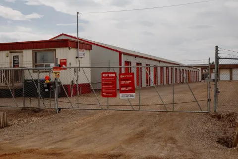 A chain link gate and red rental signs in front of a long self-storage building with red trim