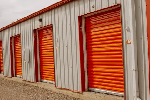 A row of outdoor drive-up self-storage units with orange roll-up doors and numbered tan metal siding
