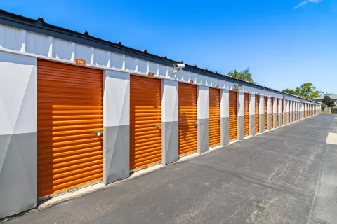 A long row of outdoor drive-up storage units with orange roll-up doors and a paved driveway