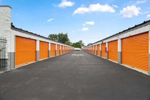 Rows of orange drive-up storage units along an asphalt driveway under a blue sky with clouds