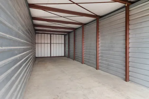 An empty indoor self-storage unit with corrugated metal walls, concrete flooring, and an overhead support structure