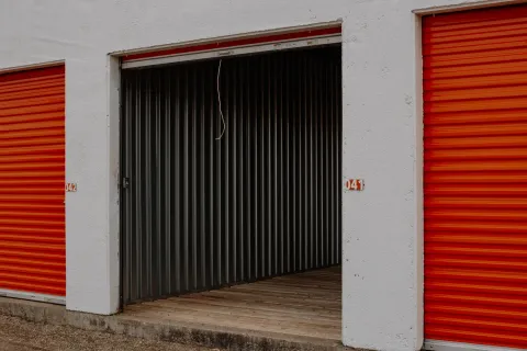 An open drive-up storage unit with a wooden floor between two closed orange roll-up doors