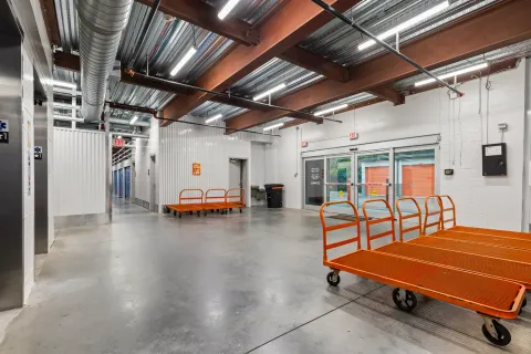 Indoor loading area with orange moving carts, glass sliding doors, and rows of storage units behind them