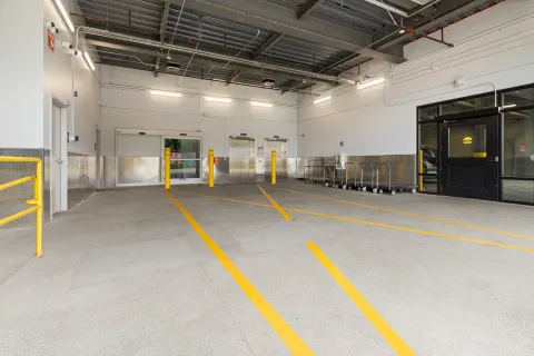 Indoor loading bay with elevators, sliding glass doors, yellow bollards, and rows of flatbed moving carts