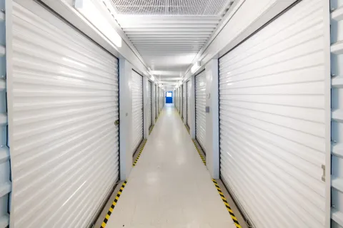 Brightly lit indoor self-storage hallway with white roll-up doors and yellow hazard floor tape leading toward a blue door