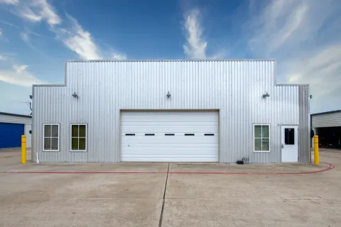 Corrugated metal storage building exterior with a white roll-up door, side windows, and a paved driveway