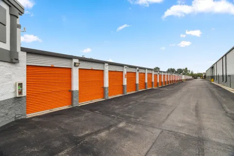 Row of outdoor drive-up storage units with orange roll-up doors along an asphalt driveway under a blue sky