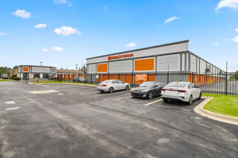 Exterior of Mini Mall Storage building with orange accents, fenced parking lot, and parked cars under blue sky
