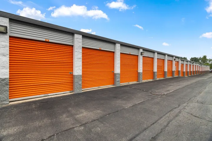 Row of drive-up self-storage units with orange roll-up doors and asphalt access path under a blue sky