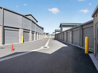 Paved drive aisle between outdoor self-storage buildings with gray roll-up doors and yellow bollards under a blue sky