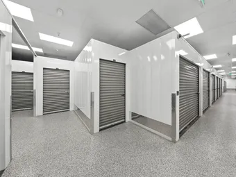 Brightly lit indoor hallway featuring white walls, gray roll-up storage unit doors, and speckled flooring