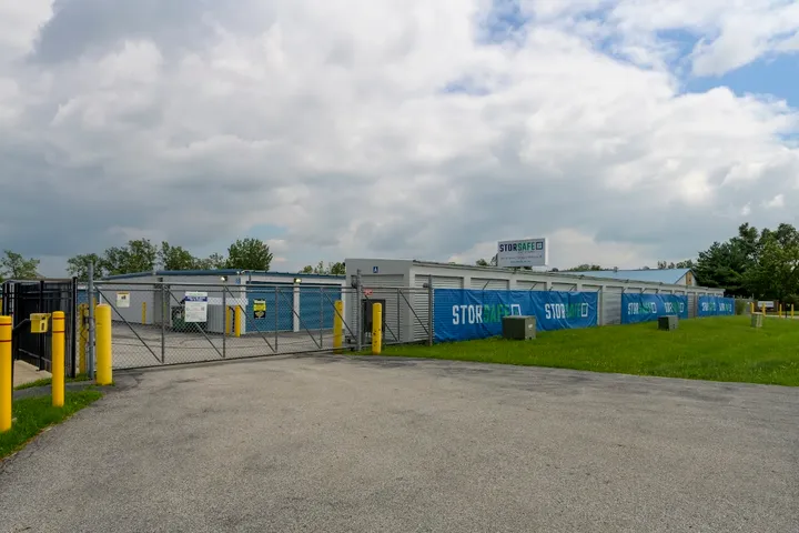 A metal gate leading to drive-up storage units with blue StorSafe banners and yellow bollards