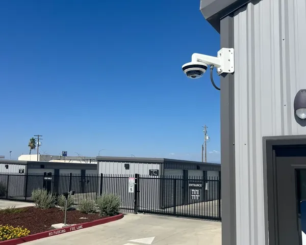 A white security camera mounted on a gray building overlooking a gated self-storage facility entrance
