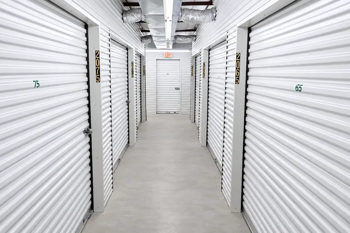 A well-lit indoor storage hallway with white corrugated unit doors, number signs, and ceiling air ducts