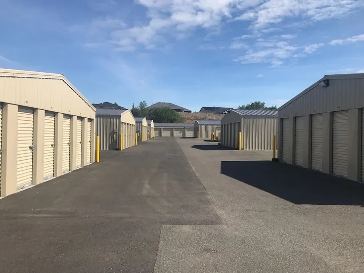 Rows of tan drive-up self-storage units with roll-up doors along a paved asphalt driveway under blue skies