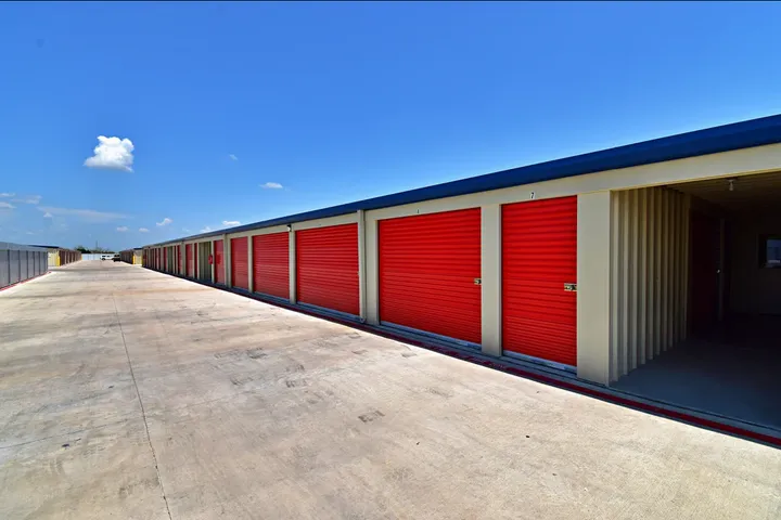 Outdoor drive-up self storage units with red roll-up doors along a wide concrete driveway under a blue sky