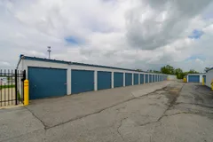 A row of drive-up storage units with blue roll-up doors along a paved asphalt driveway
