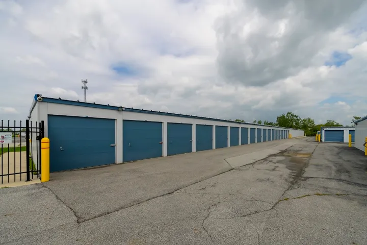 A row of drive-up storage units with blue roll-up doors along a paved asphalt driveway