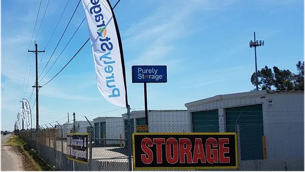 Outdoor self-storage units behind a chain-link fence with a Purely Storage sign and white feather flags