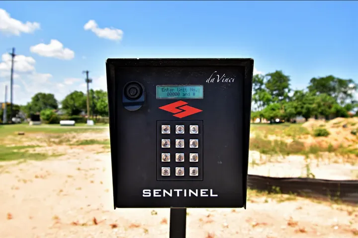 A black Sentinel gate keypad with a digital display and numbered buttons standing outdoors under a blue sky