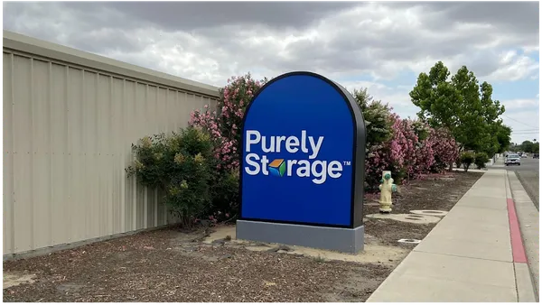 A blue arched Purely Storage sign next to a tan building and pink flowering bushes