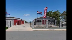 Gated entrance to a self-storage facility with red unit doors, an office building, and flags outdoors