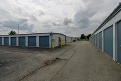 Drive-up self-storage units with blue roll-up doors and a paved driveway under a cloudy sky