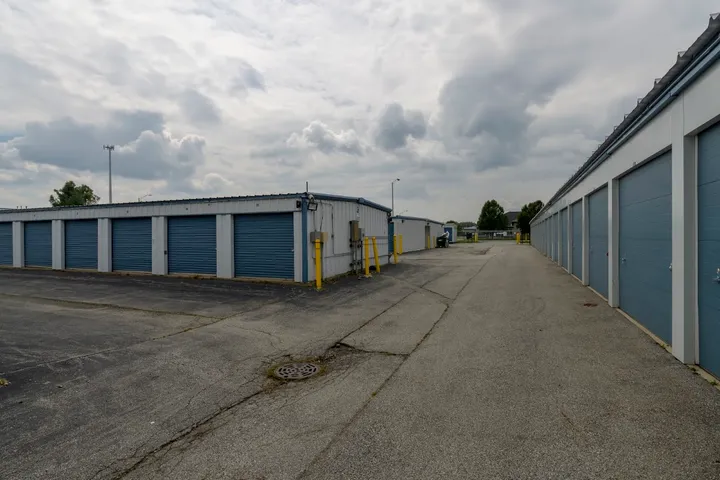 Drive-up self-storage units with blue roll-up doors and a paved driveway under a cloudy sky