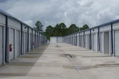 Outdoor self-storage units with white roll-up doors and blue trim along a wide concrete driving aisle