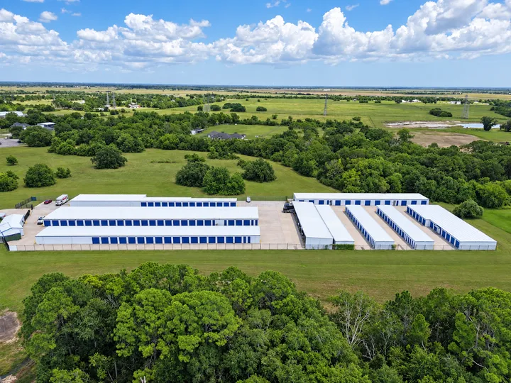 Aerial view of a self-storage facility with white roofs and blue doors surrounded by green fields