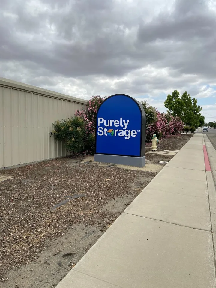 Blue Purely Storage monument sign next to pink flowering bushes and a tan metal facility wall