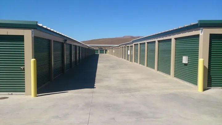 A row of outdoor drive-up storage units with green roll-up doors and yellow bollards under a clear sky