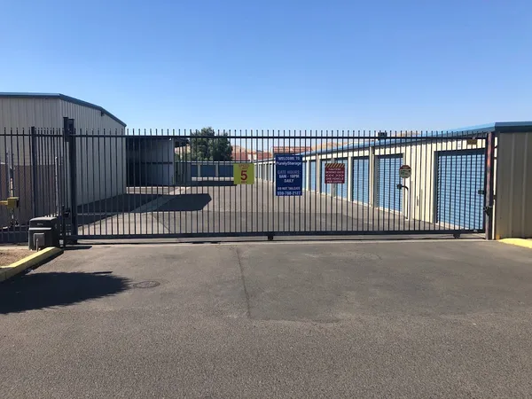 Gated entrance to a storage facility with tan buildings, blue roll-up doors, and several warning signs
