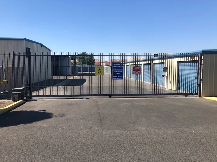 Gated entrance to a storage facility with tan buildings, blue roll-up doors, and several warning signs