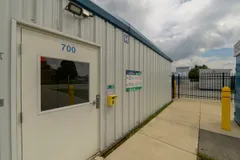 White storage facility office door with a keypad beside a metal fence and yellow bollards