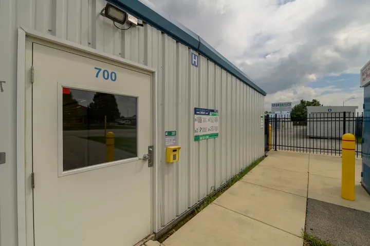 White storage facility office door with a keypad beside a metal fence and yellow bollards