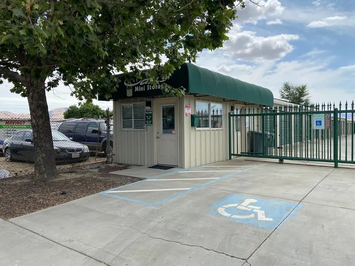 Storage facility office with a green awning, sliding metal gate, and accessible parking space under a tree