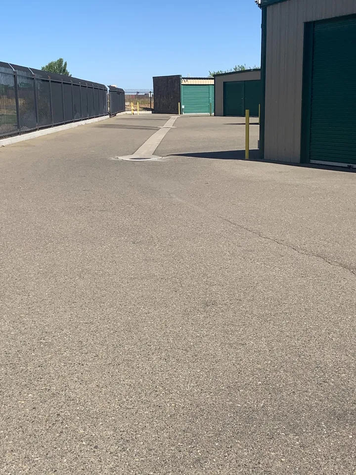 Paved drive aisle beside tan storage buildings with green roll-up doors and a black security fence