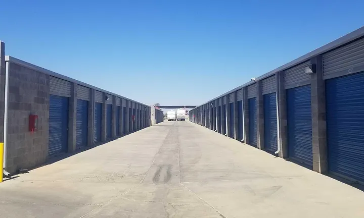Outdoor storage facility aisle with blue roll-up doors and concrete driveways under a clear blue sky