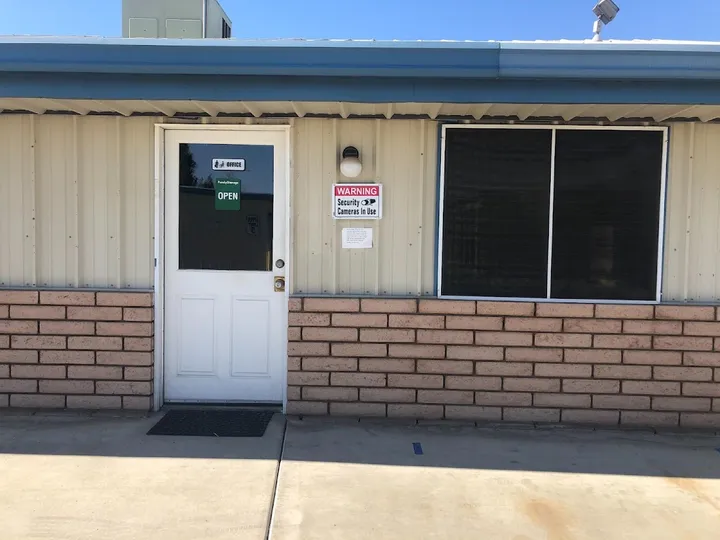 Storage facility office exterior with a white door, security camera sign, and tan metal siding