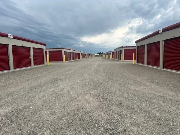 Rows of outdoor storage units with red roll-up doors along a wide gravel driving lane under cloudy skies