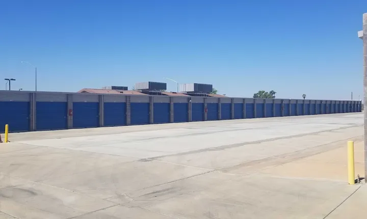 Long row of outdoor drive-up storage units with blue roll-up doors and a wide concrete driving aisle