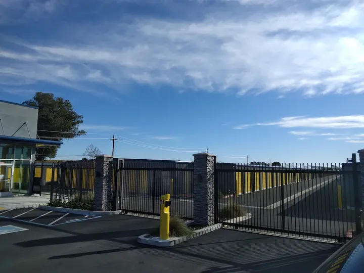 Entrance gate to the storage facility with fence and yellow unit doors visible.