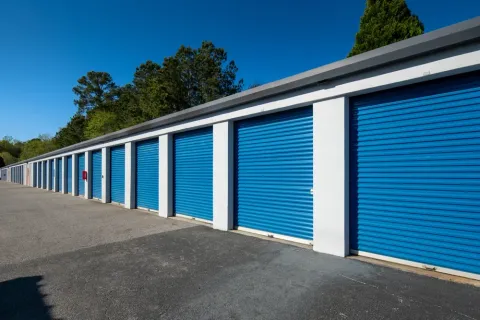 Long row of outdoor drive-up storage units with blue roll-up doors and white exterior walls