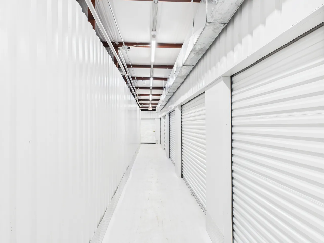 Brightly lit indoor storage hallway with white corrugated metal walls and several roll-up unit doors