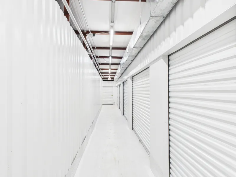 Brightly lit indoor storage hallway with white corrugated metal walls and several roll-up unit doors