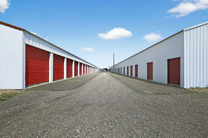 Paved driveway between two rows of white outdoor drive-up storage units in Laramie, with red roll-up doors under a blue sky