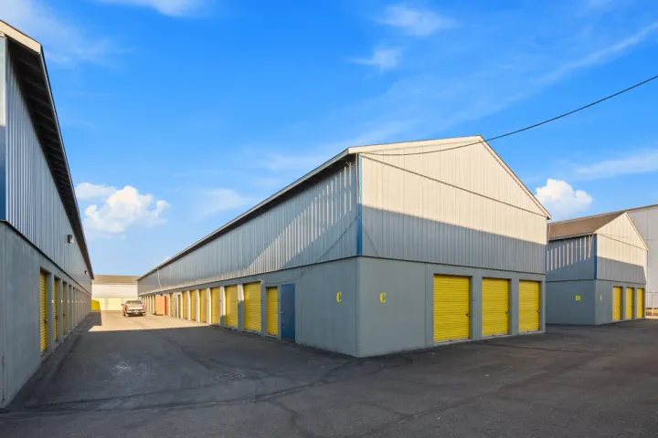 Multiple rows of outdoor storage buildings with yellow roll-up doors and asphalt driving lanes under blue sky