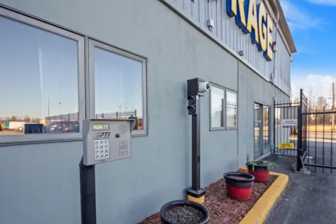 An electronic access keypad and security camera stand outside a facility building with a gated entrance