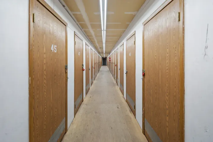 A long indoor hallway lined with numbered wooden storage unit doors under bright overhead fluorescent lighting